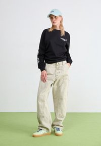 Ung kvinna med ljusblå keps, svart långärmad tröja, lösa beige byxor och gröna sneakers stående på grön golv mot vit vägg.