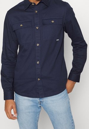 Homme portant une chemise bleu marine à manches longues avec deux poches poitrine et un jean bleu clair, main dans la poche sur fond uni.