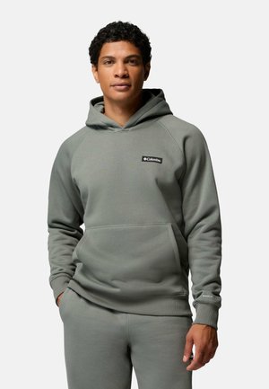 Mann trägt einen grauen Columbia-Hoodie und passende Jogginghose, steht mit einer Hand in der Tasche vor schlichtem hellem Hintergrund.