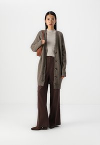 Cardigan tricotat de culoare verde închis, cu nasturi pe față, purtat peste un top cu dungi ușoare și pantaloni largi maro, asortat cu o geantă maro.
