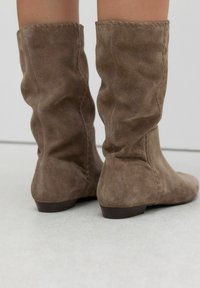Brown suede knee-high boots with a slouched design, featuring visible stitching and a low, flat heel. Textured surface with a soft finish.