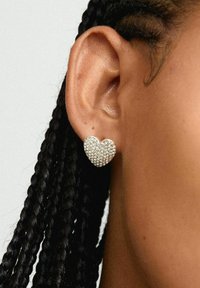Pendientes en forma de corazón hechos de plata, con una superficie cubierta de pequeños y brillantes strass, llevados en un peinado trenzado.