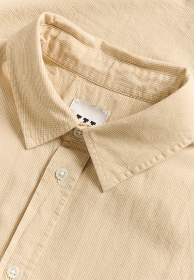 Chemise beige à col avec de fines rayures blanches, dotée d'un devant à boutons et d'une étiquette visible sous le col avec un logo.