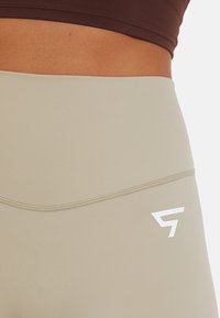 Bež legging visokog struka s glatkom teksturom, koji sadrži bijeli logo na donjoj strani i suptilni detalj šava na struku.