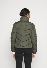 Giubbotto imbottito verde oliva con motivo a chevron, collo alto e polsini elastici. Design aderente con una texture liscia e lucida. Jeans neri indossati sotto.