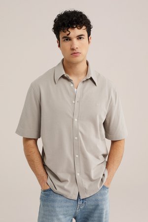 Jonge man met krullend donker haar, beige shirt met korte mouwen en lichtblauwe spijkerbroek, handen in de zakken, neutrale achtergrond.