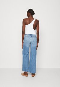 Weißes Trägershirt mit einem Schulterträger, kombiniert mit hoch taillierten hellblauen weiten Jeans. Hellbraune Slipper runden das Outfit ab.
