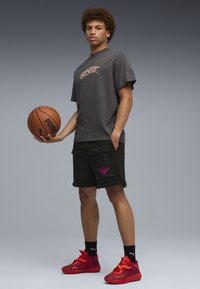 Grijs T-shirt met korte mouwen en een grafische print, zwarte, pluizige shorts met zakken, en rode sneakers met patroon. Houdt een basketbal vast.