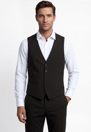 Mann trägt ein weißes Hemd mit einer schwarzen, zugeknöpften Weste und passenden schwarzen Hosen, steht mit einer Hand in der Tasche.