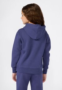 Bambino con lunghi capelli ricci indossa una felpa blu e pantaloni abbinati, di profilo su uno sfondo bianco semplice.