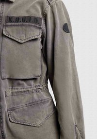 Veste grise avec deux poches à rabat, détail de taille élastique, écusson "K.H.U.J.O" sur la poitrine et patch rond noir sur la partie supérieure de la manche.