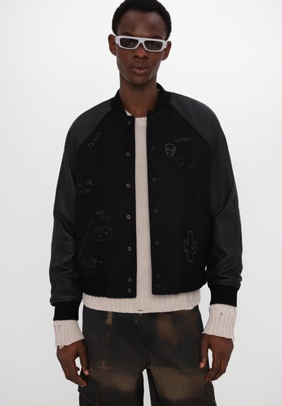 Jeune homme portant des lunettes de soleil blanches, une veste varsity noire avec des motifs brodés, un pull en tricot beige avec des poignets effilochés et un pantalon cargo marron.