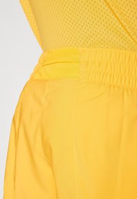 Gelbe Sportshorts aus glattem Stoff, mit elastischem Bund und seitlichen Taschen. Die Textur wirkt leicht und atmungsaktiv.