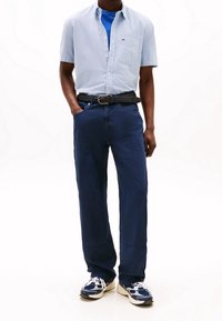 Světle modrá kostkovaná košile s krátkým rukávem přes modré tričko, doplněná tmavě modrými kalhotami a černým pleteným páskem. Výrazné bílo-modré tenisky.