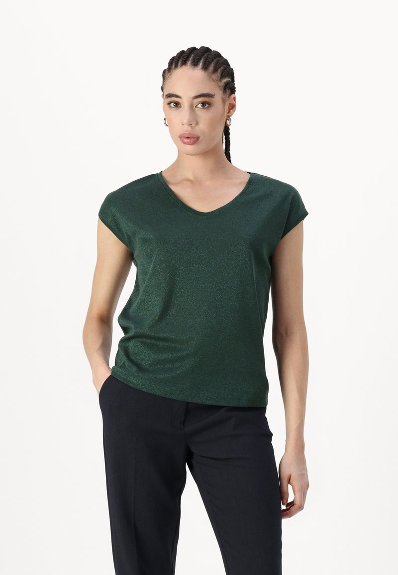 Green, short-sleeved V-neck t-shirt made of a shiny, textured fabric, paired with dark trousers, hands in pockets.
