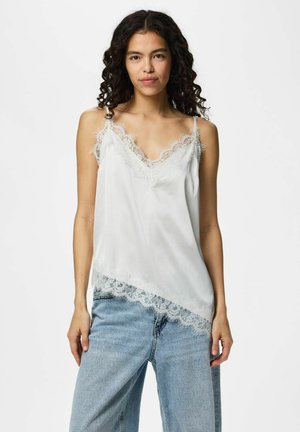 Femme aux cheveux bouclés foncés portant un caraco blanc bordé de dentelle et un jean en denim bleu clair, debout devant un fond blanc uni.