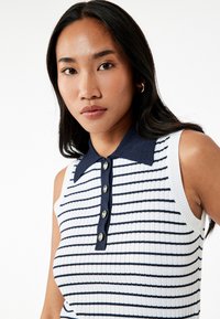Ærmeløs polo shirt i hvid og marineblå med vandrette striber. Har en ribbet tekstur, en marineblå krave og knapdetaljer.