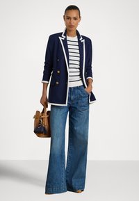 Mulher em pé, com uma mão no bolso, vestindo um blazer azul marinho com ribetes brancos, suéter às riscas, jeans de perna larga e segurando uma mala castanha.