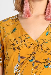 Keltainen kukkakuosinen paita, jossa V-aukko, lyhyet hihat ja napitus. Päällä yksityiskohtaiset siniset ja valkoiset kukkakuviot.