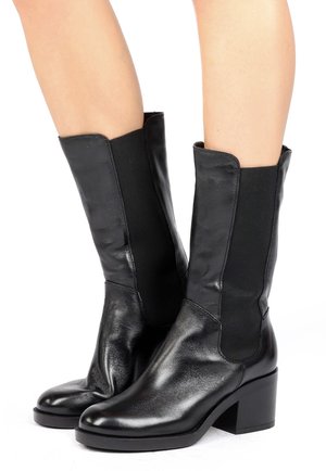 Bottes en cuir noir mi-mollet avec panneaux latéraux élastiques et talons carrés portées sur des jambes nues sur fond blanc.