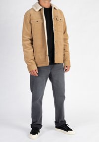 Beżowa kurtka z korduroju z pluszowym, białym kołnierzem, zapięciem na guziki, dwiema kieszeniami na piersi, noszona na czarnej koszuli i szarych dżinsach o prostym kroju.