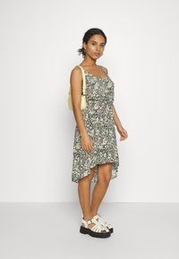 Vestido floral com fundo preto, apresentando padrões coloridos, alças finas e uma bainha com babados. Usado com sandálias brancas de tiras.