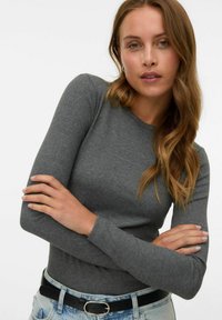 Femme aux cheveux châtain clair portant un haut gris ajusté à manches longues, un jean bleu clair et une ceinture noire, posant les bras croisés.