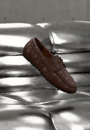 Bruine mocassinschoen met metalen veteroogjes op een zilverkleurig getuft lederen oppervlak, zwevend in de lucht alsof hij beweegt.