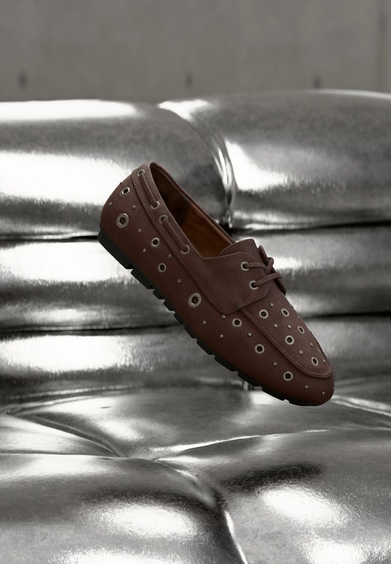 Καφετί μοκασίνι με μεταλλικά οφθαλμοθυλάκια σε ασημένια δερμάτινη επιφάνεια με ανάγλυφα, εμφανιζόμενο στον αέρα σαν να αιωρείται ή κινείται.