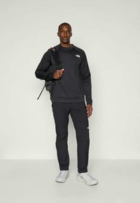 Muž stojí proti jednoduchému pozadiu, oblečený v čiernej mikine a nohaviciach North Face, s bielymi teniskami a čiernym batohom prehodeným cez jedno rameno.
