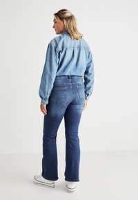 Chaqueta de mezclilla azul claro con cierre de botones, combinada con jeans acampanados azul oscuro. Zapatillas blancas completan el conjunto. Texturas suaves y diseño clásico.