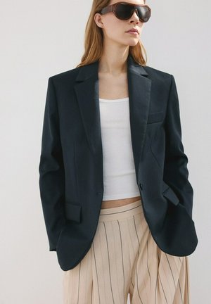 Femme portant un blazer noir oversize sur un crop top blanc, un pantalon beige à rayures fines plissé, et de grandes lunettes de soleil foncées, debout devant un fond uni.
