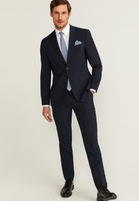 Costume bleu marine avec une veste à deux boutons et un pantalon slim. Accompagné d'une cravate bleu clair et d'un pochette. Des chaussures de ville noires complètent le look.