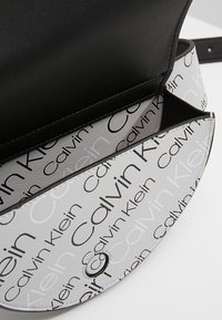 Calvin Klein-handväska i svart och vitt med en texturerad yta. Har ett upprepande logotypmönster och en cirkulär spänne för stängning.