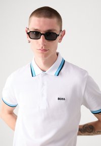 Bijela majica s okruglim izrezom i plavo-crnim prugastim ovratnikom i manžetama, s crnim logotipom "BOSS". Model nosi crne sunčane naočale, ističući detaljno vez.