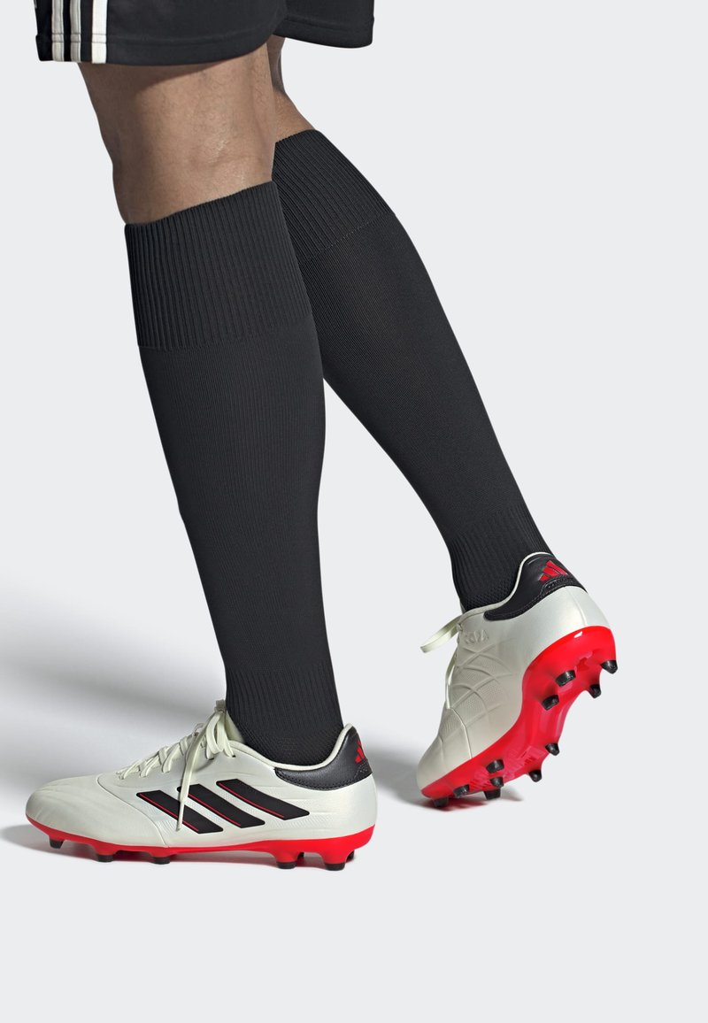 Botas de fútbol blancas con suelas rojas, franjas negras y un talón negro. Usadas con medias altas negras que muestran una textura acanalada.