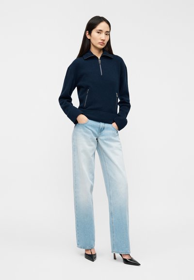 Pull marin à fermeture éclair avec deux poches latérales, associé à un jean droit bleu clair et des escarpins noirs à bout pointu. Texture lisse et design moderne.