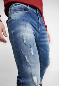 Vybledlé modré džíny z denimu se distressed detaily a trhlinami, s detailním pohledem na texturu látky a švy.