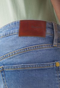 Světlé modré džínové kalhoty s hnědou koženou nášivkou s nápisem "Pepe Jeans London." Obsahují standardní zadní kapsy a žluté švy.