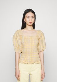 Blusa amarela xadrez com decote quadrado, corpete franzido e mangas bufantes. Usada com calças de veludo cotelê de cor clara.