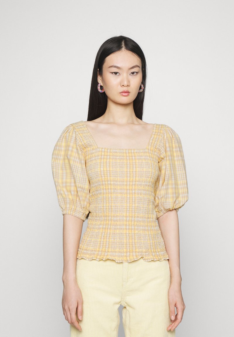 Blusa amarela xadrez com decote quadrado, corpete franzido e mangas bufantes. Usada com calças de veludo cotelê de cor clara.