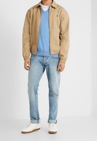 En lätt bomberjacka i tanfärg över en blå v-ringad tröja, parat med ljusblå jeans och vita sneakers med bruna sulor.