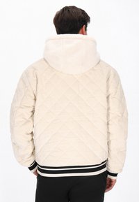 Mand iført en cremelæder quilted jakke med en fluffy hætte og sorte bukser, set fra ryggen mod en hvid baggrund.