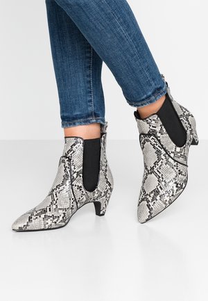 Fødder iført ankelhøje, spidse skindsko med slangeskindsdesign og sorte elastiske sider, parret med blå denimjeans.