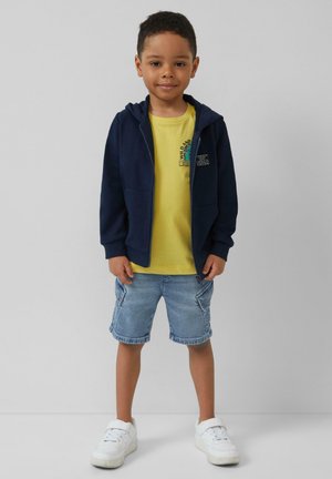 Marine rits hoodie over een gele t-shirt, gecombineerd met lichte spijkerbroekkortingen en witte sneakers; voorzien van logo's en grafische afbeeldingen op het shirt.