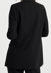 Blazer negro ajustado con hombros estructurados, solapas de muesca y un acabado suave, que presenta mangas arremangadas y una abertura trasera simple.