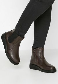 Bruna läderankelstövlar med elastiska sidopaneler, kontrasterande sömmar och låga gummisulor, visade i kombination med svarta åtsmitande byxor.