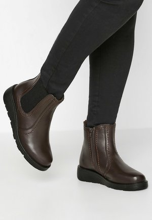 Botas de tobillo de cuero marrón con paneles laterales elásticos, costuras en contraste y suelas de goma bajas, mostradas usadas con pantalones ajustados negros.