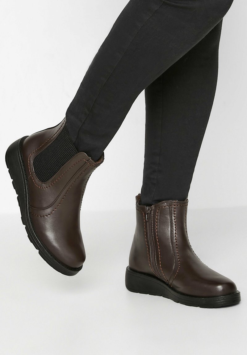 Bruna läderankelstövlar med elastiska sidopaneler, kontrasterande sömmar och låga gummisulor, visade i kombination med svarta åtsmitande byxor.