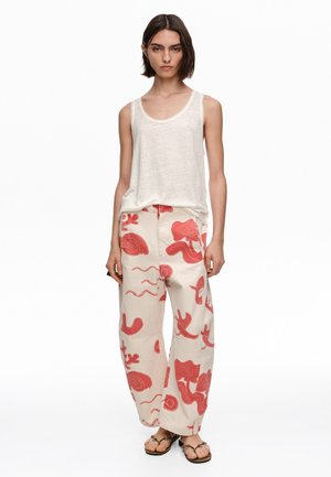 Kobieta nosząca białą bluzkę bez rękawów, szerokie kremowe spodnie z czerwonymi abstrakcyjnymi wzorami oraz płaskie sandały, stojąca na białym tle.